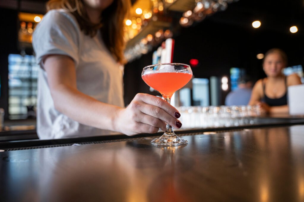Close up of a hand holding a pink signature craft cocktail in a coupe glass on the bar counter at The Bench Fort Worth