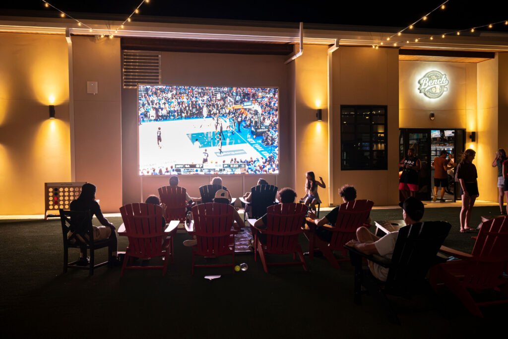 Guests watching a live basketball game on the outdoor jumbo screen with lawn chairs at The Bench sports bar patio in Fort Worth