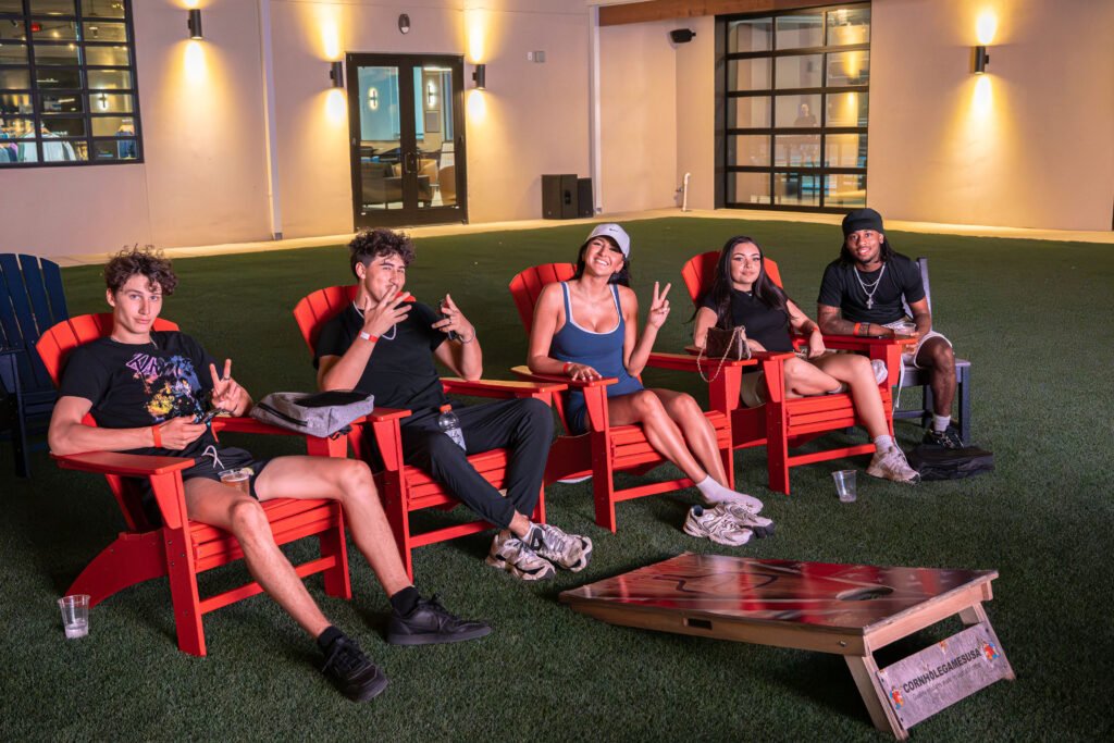 Group of friends hanging out in red Adirondack lawn chairs on the turf patio next to cornhole boards at The Bench sports bar in Fort Worth