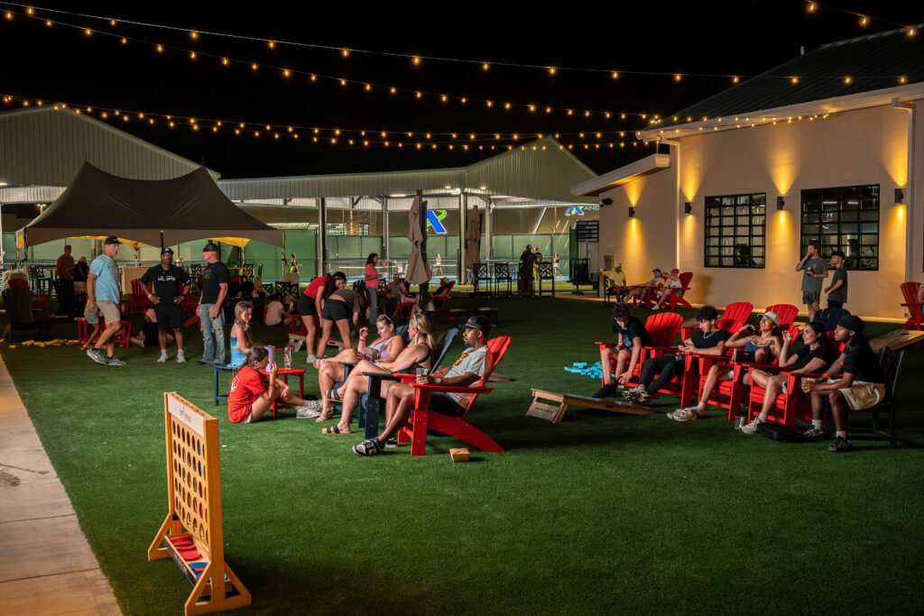 Crowd relaxing in lawn chairs on the artificial turf patio at The Bench sports bar and pickleball venue at night